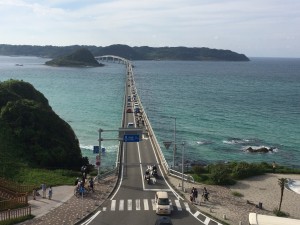 ターコイズブルーの海にかかる長い角島大橋は遠くの島へと続いており、道路や海岸には車や人が行き交っています。.
