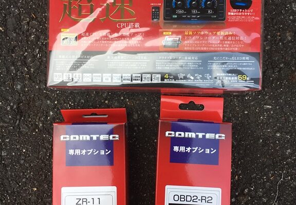 アスファルトの上に、COMTEC の車載電子機器ボックス 2 つと、大きな赤い COMTEC OBDII パッケージ 1 つがあります。.
