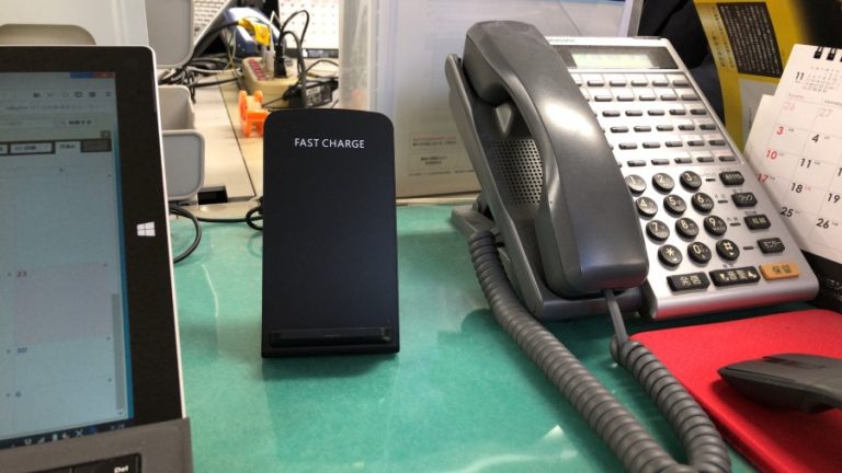 電話、FAST CHARGE というラベルの付いたワイヤレス充電器、ノートパソコンの一部が見えるオフィスデスク。.