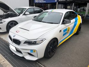 青と黄色のレーシング デカールが付いた白い BMW スポーツカーが、別の車の隣に屋外に駐車されています。.