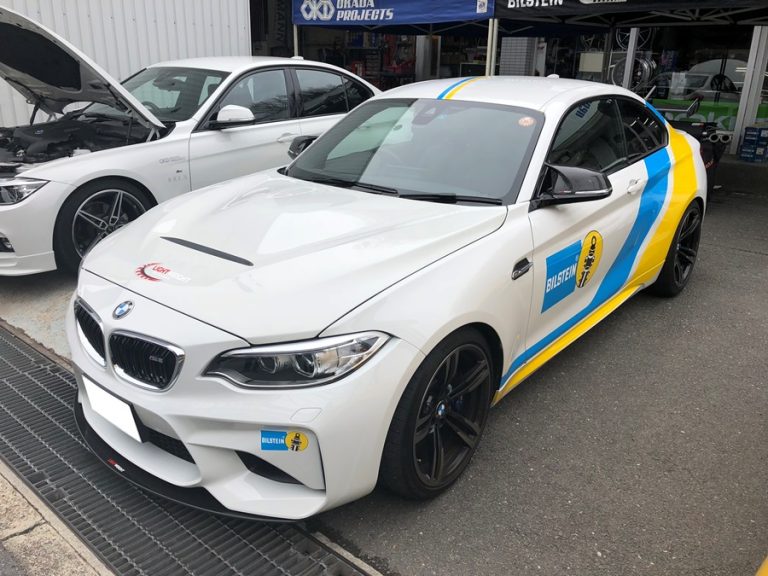 青と黄色のレーシング デカールが付いた白い BMW スポーツカーが、別の車の隣に屋外に駐車されています。.