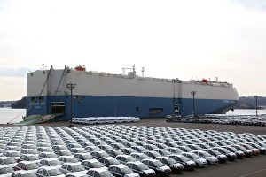 大型貨物船が港に停泊しており、前景には白い新車が何台も並んでいる。.