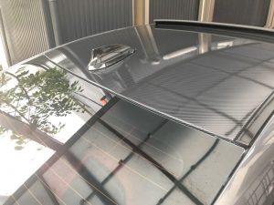 シャークフィンアンテナ、反射する木々、金属製のフェンスを備えたカーボンファイバー製の車の屋根。.