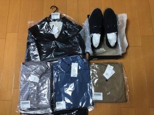 タグとパッケージが付いたシャツ、ズボン、ジャケット、黒い靴が木の床の上に並べられています。.