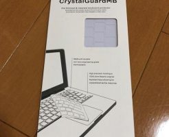 CrystalGuardMB キーボード プロテクターのパッケージが木の床に置かれ、ラップトップとシリコン カバーのグラフィックが表示されています。.