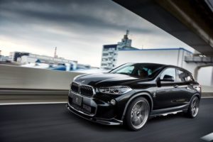 黒い BMW SUV が高架下の市街地高速道路を走行しており、背景にはぼやけた建物とドラマチックな空が広がっています。.