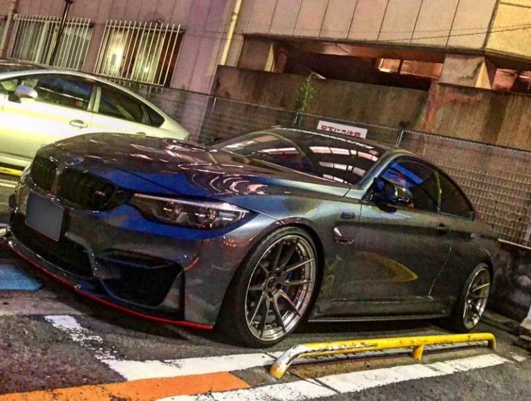 夕暮れ時の都市部の駐車場に、大きなホイールを備えた洗練されたグレーのスポーツカーが斜めに駐車されている。.