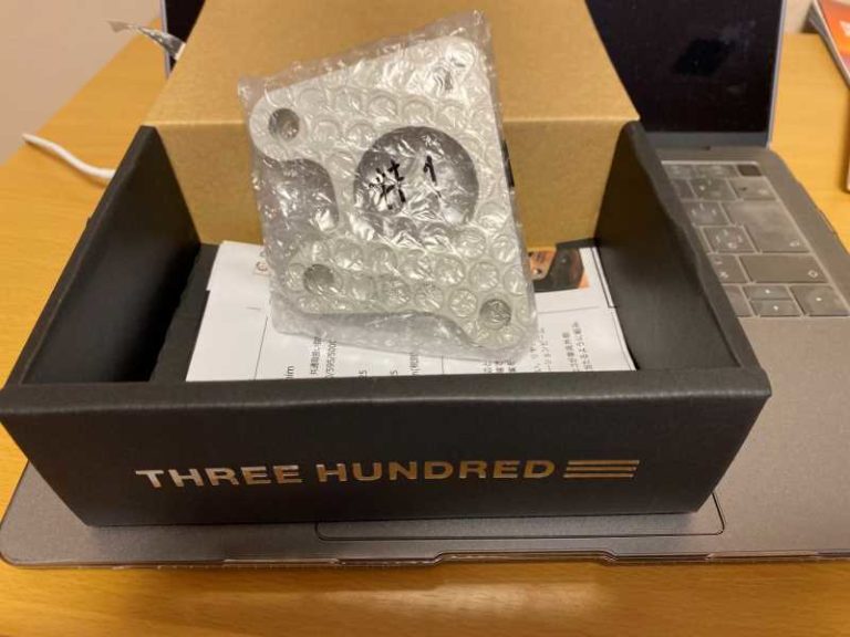 気泡緩衝材で包まれた金属部品が、ノートパソコンと木製の机の上に置かれた「THREE HUNDRED」というラベルの付いた箱の中に入っています。.