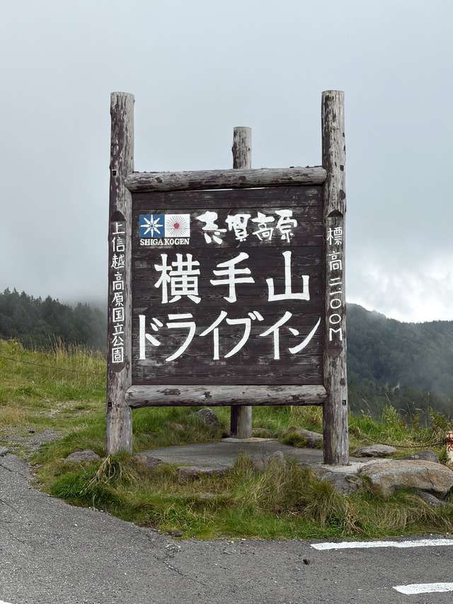 霧の山中に立つ木製看板。「横手山ドライブイン」「標高2100m」「上信越高原国立公園」と記されている。