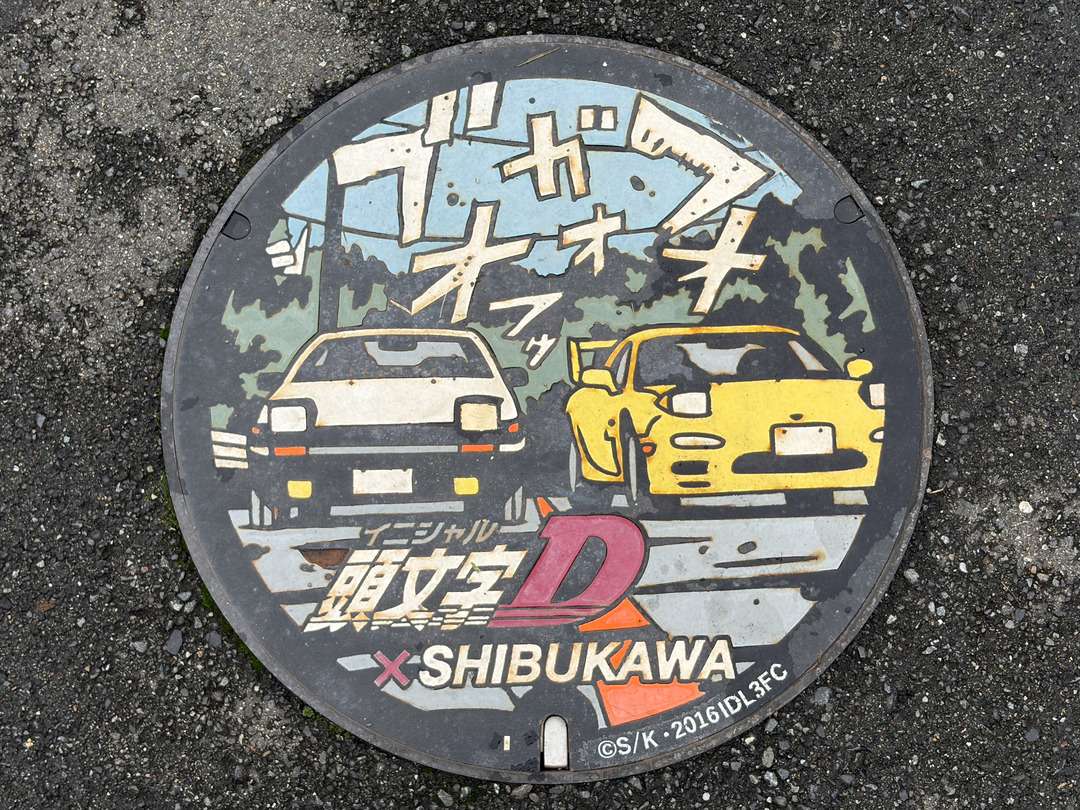 『頭文字D』のロゴと2台の車が描かれた、SHIBUKAWA(渋川)のマンホール蓋。