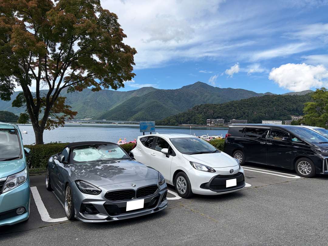 湖畔の駐車場に複数の車が並び、背後に橋と山々が広がっている。