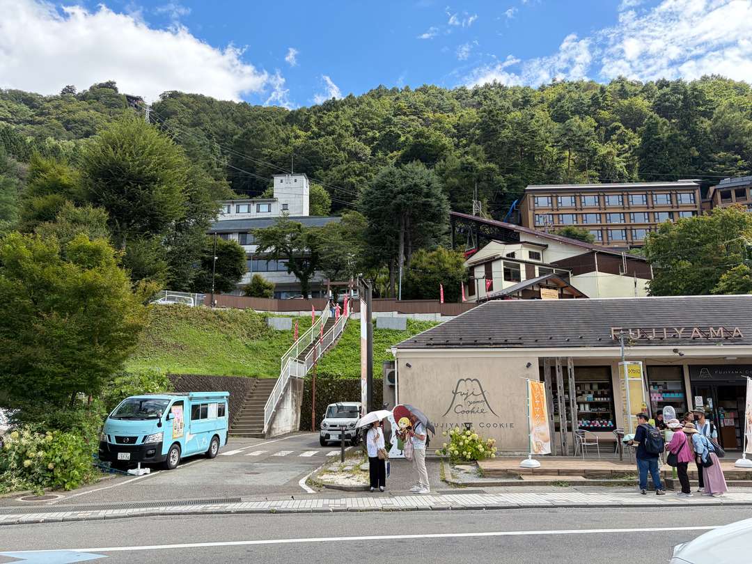 山の斜面に建つFUJIYAMA COOKIEの店舗と、前の道路を歩く観光客、青い車両が写っている。