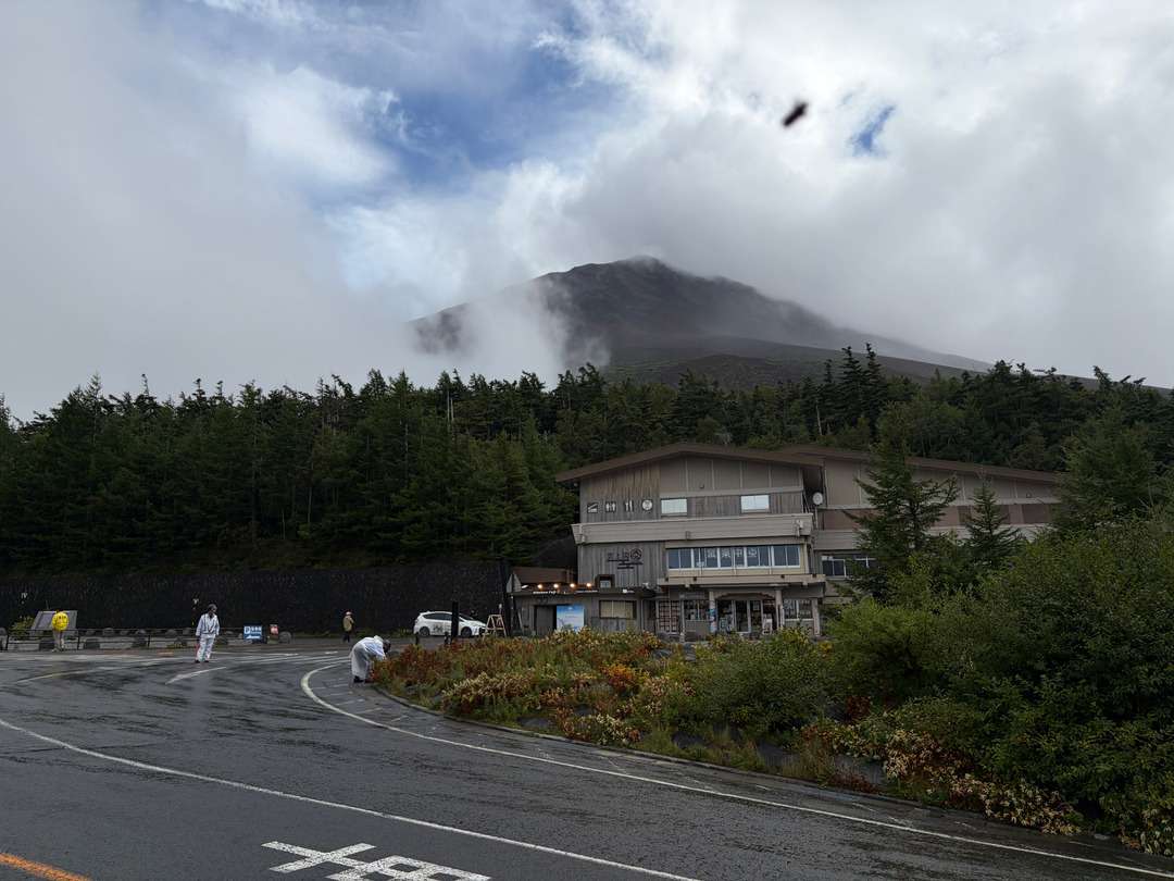 雲に覆われた山を背景に、森林の中の山間施設と雨に濡れた駐車場が写っている。
