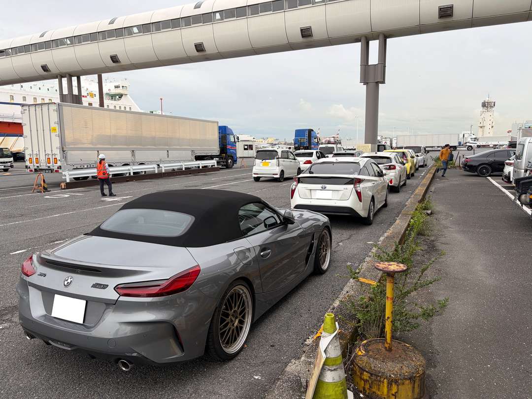 立体歩道橋の下で乗用車やトラックが並ぶ駐車場。近景に幌付きのBMWが停車している。
