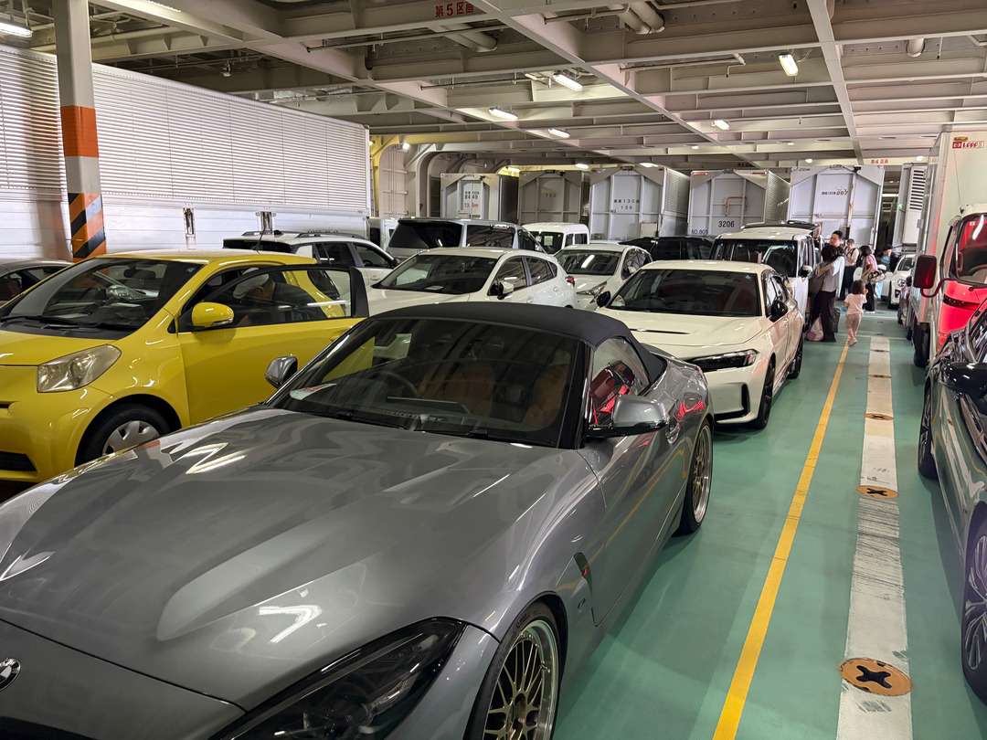 駐車場内に黄色や白、灰色などの乗用車が並び、奥にトラックと人々が見える。